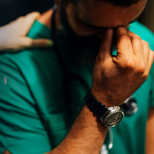 A healthcare professional in scrubs holding his hand to his face, showing signs of stress, with a supportive hand on his shoulder.