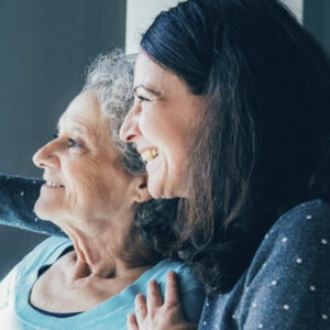 Two women smiling as they look out of a window, one older and one younger, standing close together with their arms around each other.”